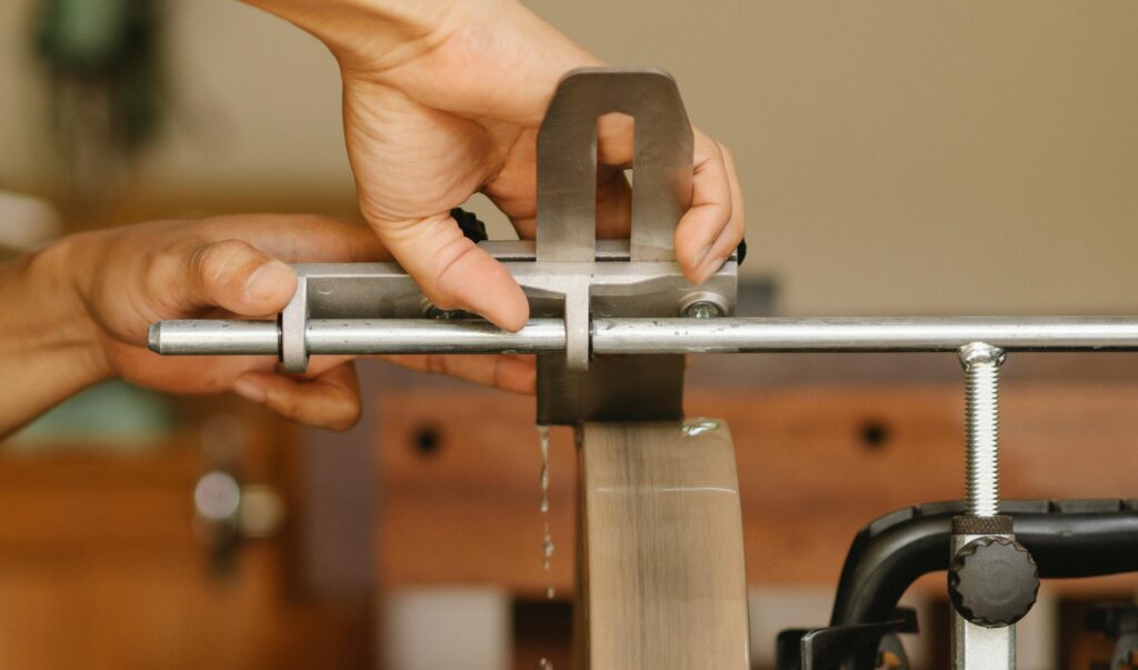 Side view of unrecognizable craftsman sitting at sharpening machine with metal stick while grinding knife in professional studio on blurred background
