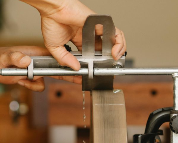 Side view of unrecognizable craftsman sitting at sharpening machine with metal stick while grinding knife in professional studio on blurred background